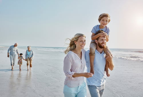 A family enjoys a sunny day on the beach. A man carries a smiling child on his shoulders, walking alongside a woman. An elderly couple and child stroll behind them.