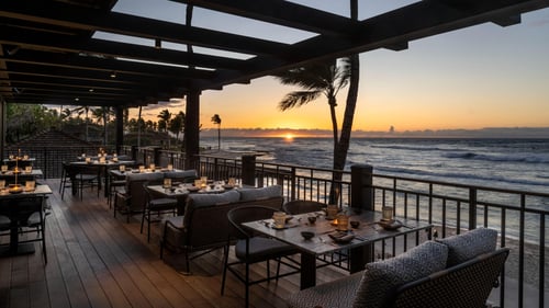 Oceanfront restaurant terrace with candlelit tables and cushioned seating overlooking a sunset, palm trees and waves