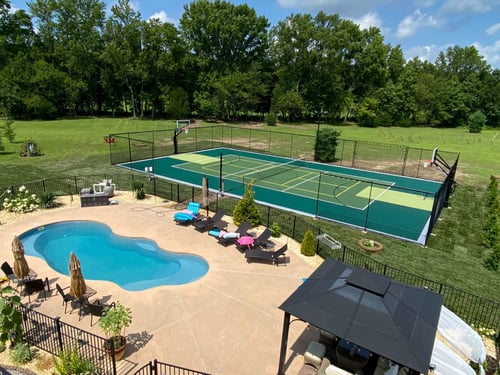 Overhead shot of a backyard pool area next to a fenced multi-purpose sports court surrounded by a lush lawn and trees.