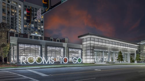 Exterior view of the Rooms To Go furniture store in Buckhead, Atlanta, GA, at dusk with illuminated signage, large windows, and the Hyatt Place hotel in the background.