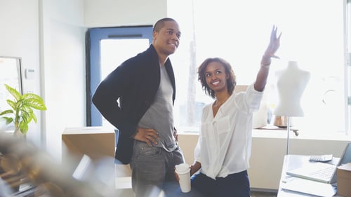 Two people in a bright space discuss ideas. A woman, smiling, gestures upward with enthusiasm, holding a coffee cup. A man stands beside her, listening intently.