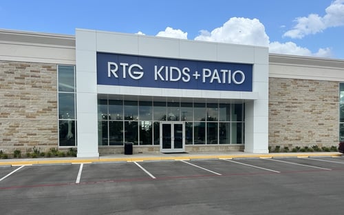 Exterior of the South Austin RTG Kids + Patio store with a modern facade, stone accents, large glass windows, and a blue sign above the entrance on a sunny day.