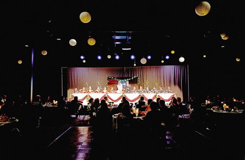 Dimly lit event hall with round banquet tables, a stage, and string lights for a formal reception.
