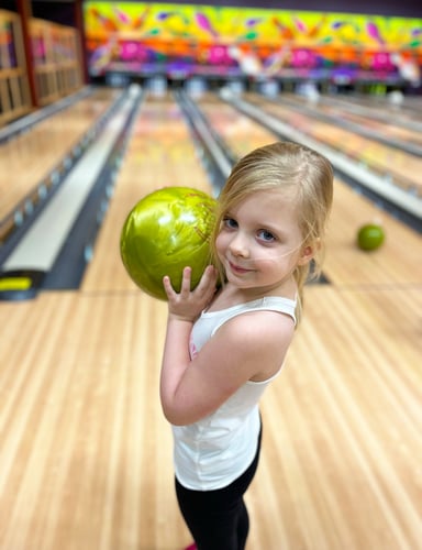Girl holds bowling ball and looks back a t camera. Bowling lane is in background,