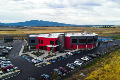 Peak Post Fall's new two-story gym has a striking red exterior and large windows. The building is surrounded by a well-maintained parking lot with ample spaces, including marked accessible spots. The gym is situated in an open area with expansive fields and mountains visible in the background.