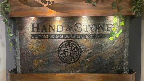 A pristine, softly lit indoor cascading stone wall fountain featuring prominent corporate branding in the luxury reception lobby of Hand & Stone Spa in Paradise Valley, Arizona.