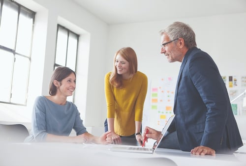 Three colleagues are engaged in a friendly discussion around a laptop in a bright office. The atmosphere is collaborative and positive.