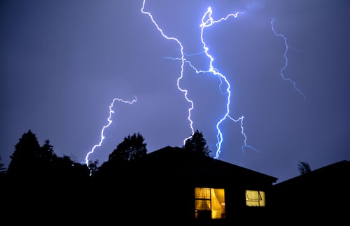 Lightning lights up the sky behind a home with lights coming through the windows.