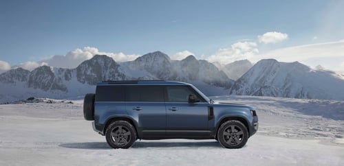 Side view of a blue Defender 110 parked on a snowy mountain landscape with rugged peaks in the background
