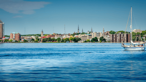 Sailboat on calm blue water with a vibrant city skyline in the background, featuring mix of modern and historic buildings under a clear sky.