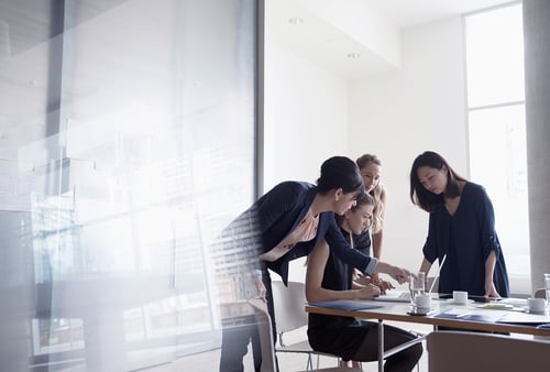 Four women collaborating at a table in a bright office, focused on paperwork and a laptop, conveying teamwork and concentration.