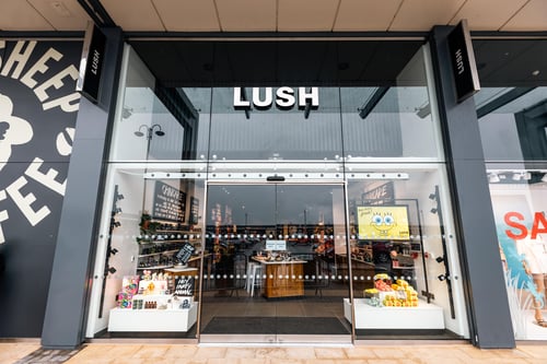 Exterior view of shop. Black framed shop with glass frontage. Large door way in centre with illuminated Lush sign above. Products displayed in windows either side of entrance.