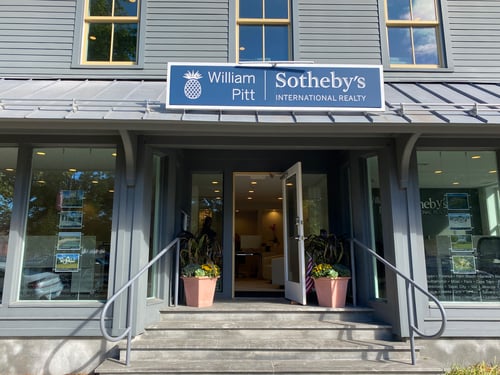 Gray building with metal awning and glass storefront housing William Pitt Sotheby's International Realty in Guilford, Connecticut, featuring open entrance door with curved railings and flower planters.