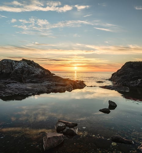 Serene sunset over the ocean, framed by dark rocky cliffs. The sky reflects vibrant hues of orange and pink in the calm water below.