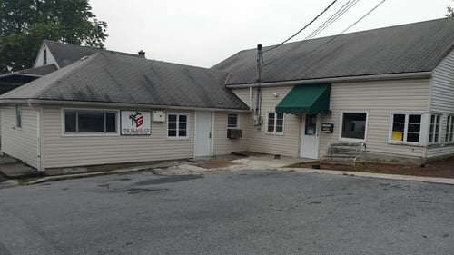 The attached image shows the exterior of a small, beige building that houses the “ITG Glass Company.” The building has a simple, low-profile design with a gray shingled roof and siding. A sign with the “ITG Glass Company” logo is mounted on the wall near the entrance, which is a white door with a small window. The entrance area also features a green awning above another door, adding a touch of color to the otherwise neutral facade. The building is situated on a quiet, slightly sloped street, and there is a metal bench near the green awning. The overall setting appears modest and functional, reflecting a straightforward, professional business environment.