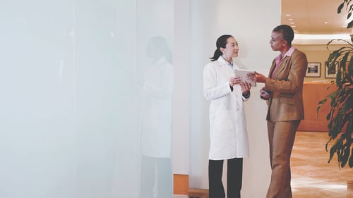 A doctor in a white coat and a businesswoman in a brown suit are engaged in a conversation in a bright, modern office. The mood is professional and collaborative.