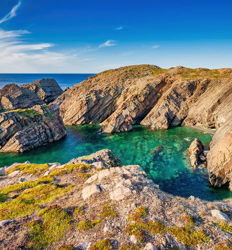 Rocky cliffs surround a turquoise inlet under a clear blue sky. The vibrant greenery and rugged terrain add contrast and tranquility to the scene.