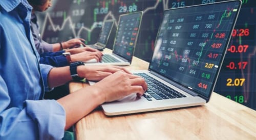 Hands typing on a laptop computer that displays stock market data on the screen.