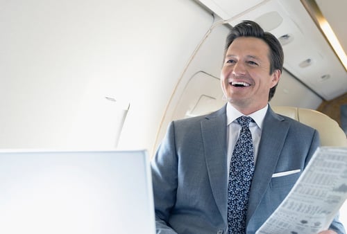 Smiling man in a suit holding a newspaper, seated in a well-lit airplane. He appears relaxed and content, embodying a professional and confident tone.