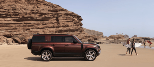 Side view of a red Defender 130 parked on a sandy beach with surfers nearby.
