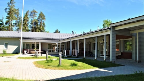 Ulkokuva Pellaksesta. Rakennus on yksikerroksinen ja vaalean värinen. Pihalla on kiveystä, nurmikkoa ja pihatiet on valaistu kunnolla. Taivas on sininen ja talon takana näkyy metsää.