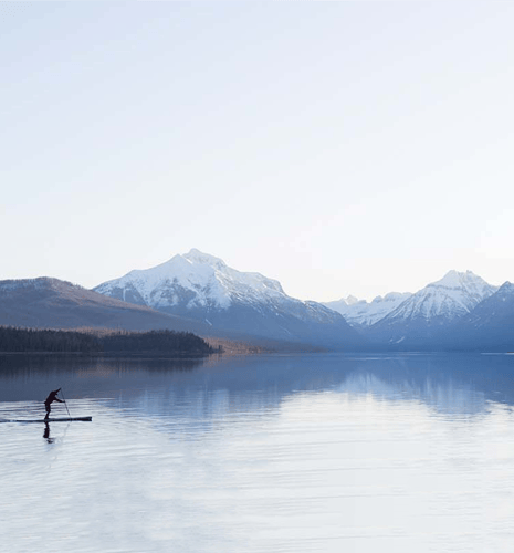 A person in red paddleboarding on a calm lake, surrounded by snow-capped mountains under a clear blue sky, conveying serenity and adventure.