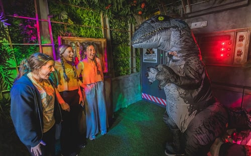 Three excited players encounter a person in an inflatable T-Rex costume, experiencing a fun scare in the Raptor Revenge room at OBXscape Rooms