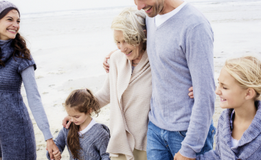 A happy multigenerational family walks on a beach, dressed in cozy sweaters. They smile warmly, conveying togetherness and joy. Overcast sky in the background.