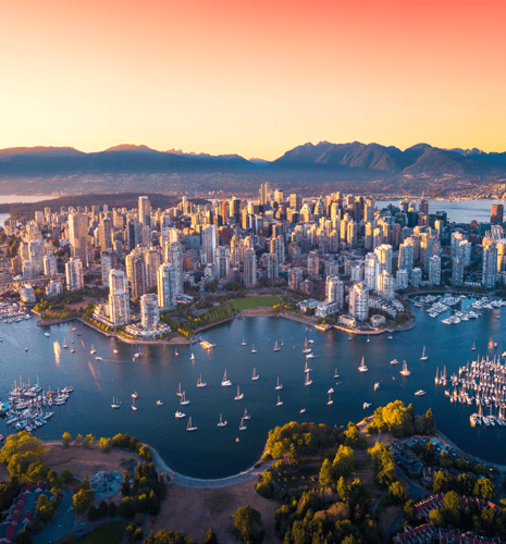 Aerial view of Vancouver at sunset with skyscrapers, mountains in the background, and a harbor filled with sailboats. The sky is a warm orange.