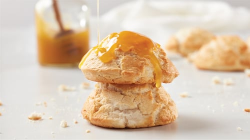 Stacked golden biscuits with honey sauce being drizzled on top, surrounded by extra biscuits and a jar of honey sauce in the background on a crumb-covered surface.