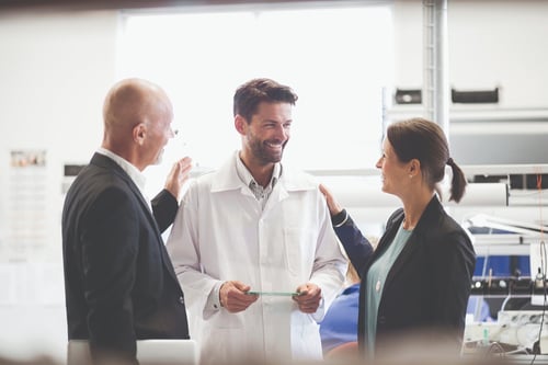 Three people in a lab setting, two in business attire congratulating a smiling man in a lab coat, suggesting teamwork and success.