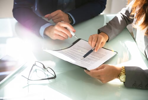 Two business professionals review and sign a document at a glass table with glasses and a pen nearby.