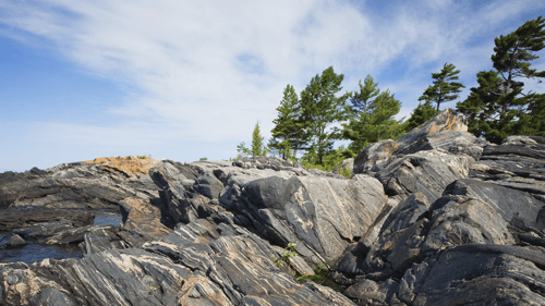 Rocky shoreline with rugged, layered stones and scattered green coniferous trees under a bright blue sky with wispy clouds. Serene and natural.