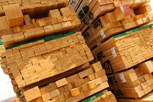 Stacks of neatly arranged lumber are piled high in an outdoor storage area. The wooden boards are cut uniformly, with ends facing outward, showcasing the natural grain and texture of the wood. Some of the stacks are bound with green straps, and several boards are marked with barcoded labels and black spray-painted numbers.