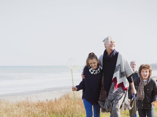 A joyful family strolls along a grassy beach path, with the ocean in the background. A young girl holds a fishing net, smiling alongside an older woman.