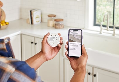 Man holding a water leak/freeze detector and a smart phone showing the Kidde app, standing in front of a kitchen sink.