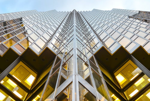 Upward-looking view of a modern glass and steel skyscraper shot from ground level. The building features a symmetrical geometric design with a sharp central peak, reflective glass panels, and a grid of illuminated yellow-tinted windows. The facade creates a striking vanishing point perspective leading toward the sky, with metal framework and architectural lines emphasizing the building's height and contemporary design.