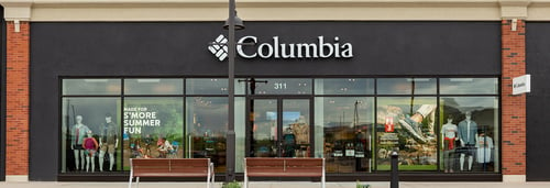 Photograph of Columbia Sportswear storefront with black and white logo sign above the entrance. The large glass windows display mannequins dressed in outdoor clothing, and the store's interior is visible. The suite number 311 is shown above the door. Two wooden benches are placed outside the store on either side of the entrance.