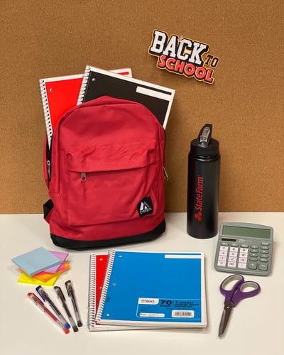 A red backpack surrounded by notebooks, pens, scissors, a calculator, sticky notes, and a water bottle on a table, with a "Back to School" sign.