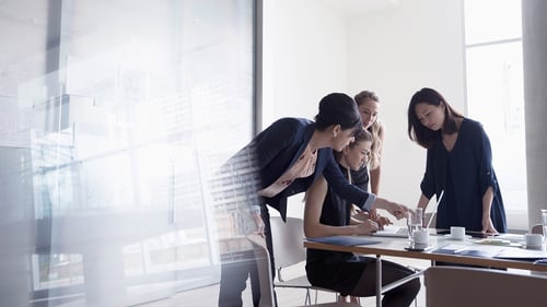 Four women collaborating at a table in a bright office, focused on paperwork and a laptop, conveying teamwork and concentration.