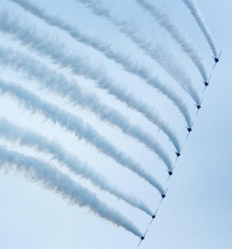 Nine jet planes fly in formation against a clear blue sky, leaving thick, curved trails of white smoke. The scene conveys precision and unity.