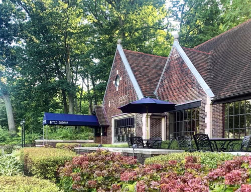 Red brick building with steep gabled roofs and blue awning housing Julia. B Fee Sotheby's International Realty in Scarsdale, New York, featuring outdoor seating patio and colorful flower gardens.