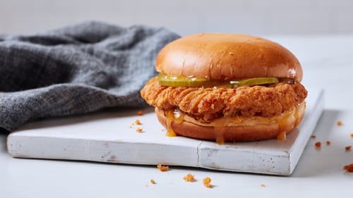 This image showcases a delicious fried chicken sandwich. The crispy chicken breast is tucked between a soft, lightly toasted bun with pickles adding a burst of freshness. The sandwich is served on a clean white plate, with some crumbs scattered around, emphasizing its crispy texture. A cozy gray cloth in the background adds a warm, rustic touch to the overall presentation.