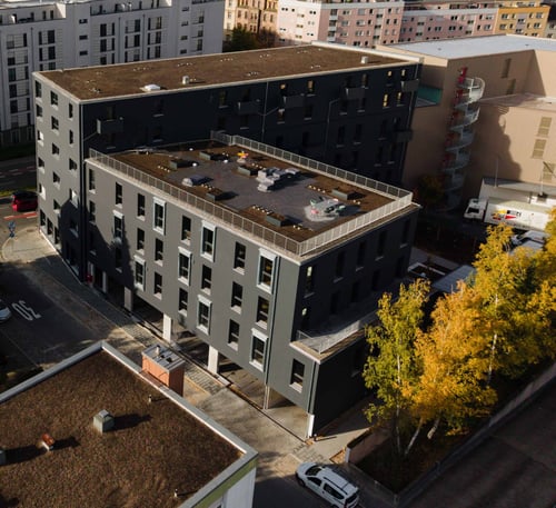 Modernes Apartmenthaus für Geschäftsreisende in zentraler Stadtlage, dunkle Fassade mit Flachdach und großzügiger Dachterrasse mit Sitzbereichen, umgeben von weiteren Büro- und Wohngebäuden sowie einer Straße mit parkenden Autos, im Vordergrund Bäume mit herbstlich gelben Blättern.