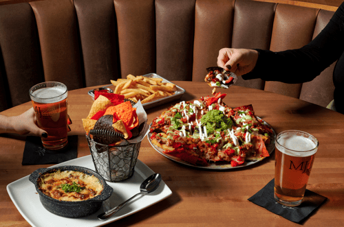 A table filled with shareable appetizers at MJ23 Sports Bar & Grill, featuring loaded nachos with guacamole, a bubbling cheese dip with tortilla chips, crispy fries, and cold pints of beer.