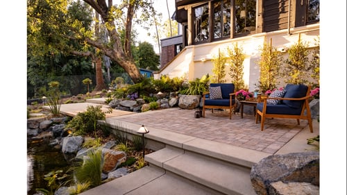Serene landscaped patio with two blue armchairs beside a pond, stone steps, lush garden, and sunlit house; a welcoming outdoor retreat.