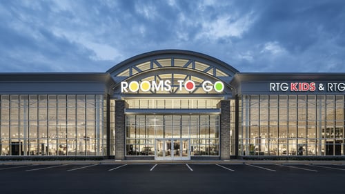 Front view of Rooms To Go store in Virginia Beach, VA, at dusk with expansive glass windows and illuminated interior.