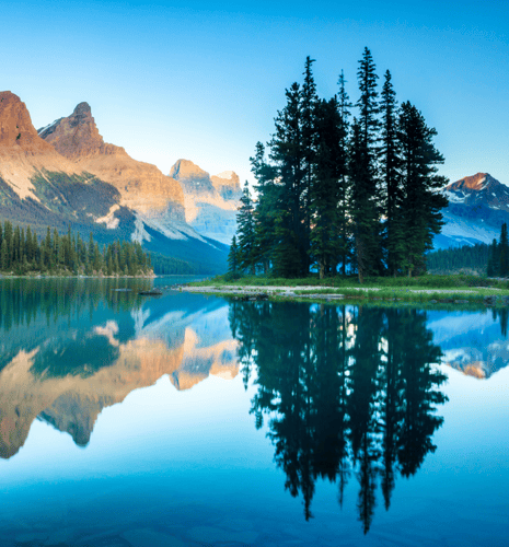 Serene lakeside scene at sunrise, with evergreen trees on an island reflecting in calm water. Majestic mountains bathed in golden light in the background.