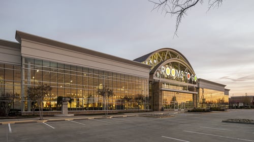Exterior of a Rooms To Go Kids store at sunset, showcasing expansive glass walls reflecting warm light and a spacious parking area.