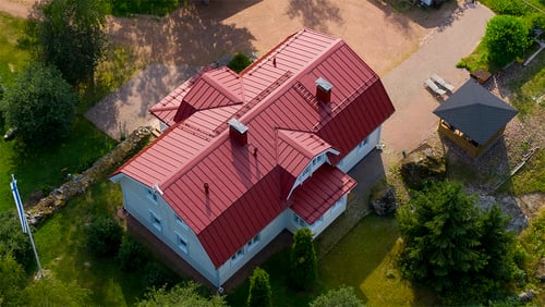 Kuvassa on ilmasta otettu valokuva suuresta kaksikerroksisesta puutalosta, jossa on punainen mansardikaatto. Vesikatemateriaali on Ruukki Classic. Talon katossa on useita harjanteita ja kulmia jotka heijastavat valoa kauniisti. Talon seinät ovat vaaleat ja talossa on valkoiset ikkunanpuitteet. Talon ympärillä on hyvin hoidettu piha, jossa näkyy erilaisia puita ja pensaita, ja se rajautuu siistiin nurmikenttään. Pihaan kuuluu myös kivetty ajotie. Kuvassa on auringonvalon ja varjojen leikki, mikä korostaa rakennuksen ja sen ympäristön yksityiskohtia.